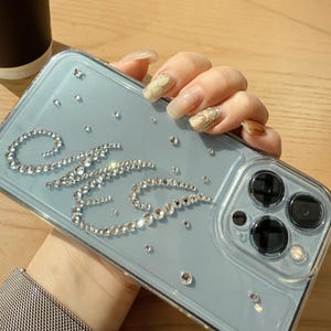 May include: A clear phone case with a light blue backing, adorned with sparkling rhinestones spelling out the letters "ME". Additional scattered rhinestones add to the case's decorative appeal. The case is held by a person.