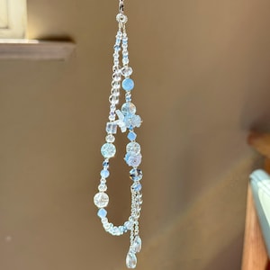 Pu&ograve; includere: Un ornamento di perline fatto a mano con perline trasparenti, azzurre e argentate. L'ornamento include perline rotonde, a goccia e a forma di fiore, sospese da un sottile cordoncino color argento. Il design ha una qualit&agrave; stravagante ed eterea.