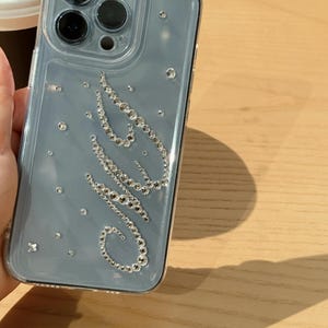 May include: A clear phone case with a light blue backing and sparkling rhinestone embellishments. The rhinestones form the word "OMG" in cursive script, with additional scattered rhinestones. The case has a clear border and covers the phone's camera.