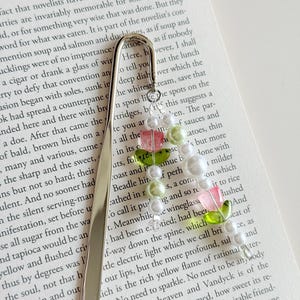May include: A decorative silver bookmark featuring a beaded floral design. The beads are in shades of white, pink, green, and clear. The bookmark is placed on the pages of a book, adding a touch of elegance.
