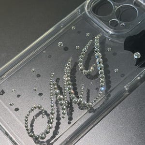 May include: Clear phone case with the letters "MJ" spelled out in silver rhinestones. The case has a clear, transparent design with scattered rhinestones throughout. The case is designed to fit a phone.