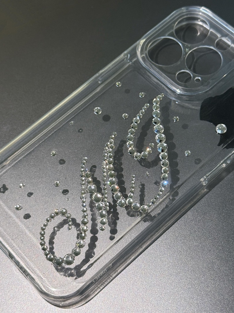 May include: Clear phone case with the letters "MJ" spelled out in silver rhinestones. The case has a clear, transparent design with scattered rhinestones throughout. The case is designed to fit a phone.