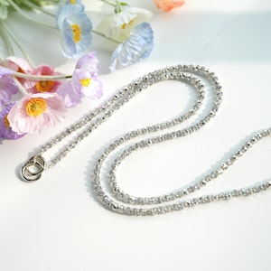 May include: A delicate silver-colored beaded necklace with a double-loop clasp. The necklace is draped on a white surface, with colorful poppy flowers in the background. The flowers have purple, pink, and blue petals with yellow centers.