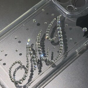May include: Clear phone case with the word "NYC" spelled out in sparkling rhinestones. The case has a transparent design with scattered rhinestones, and is designed for a phone. The case is made of clear plastic.