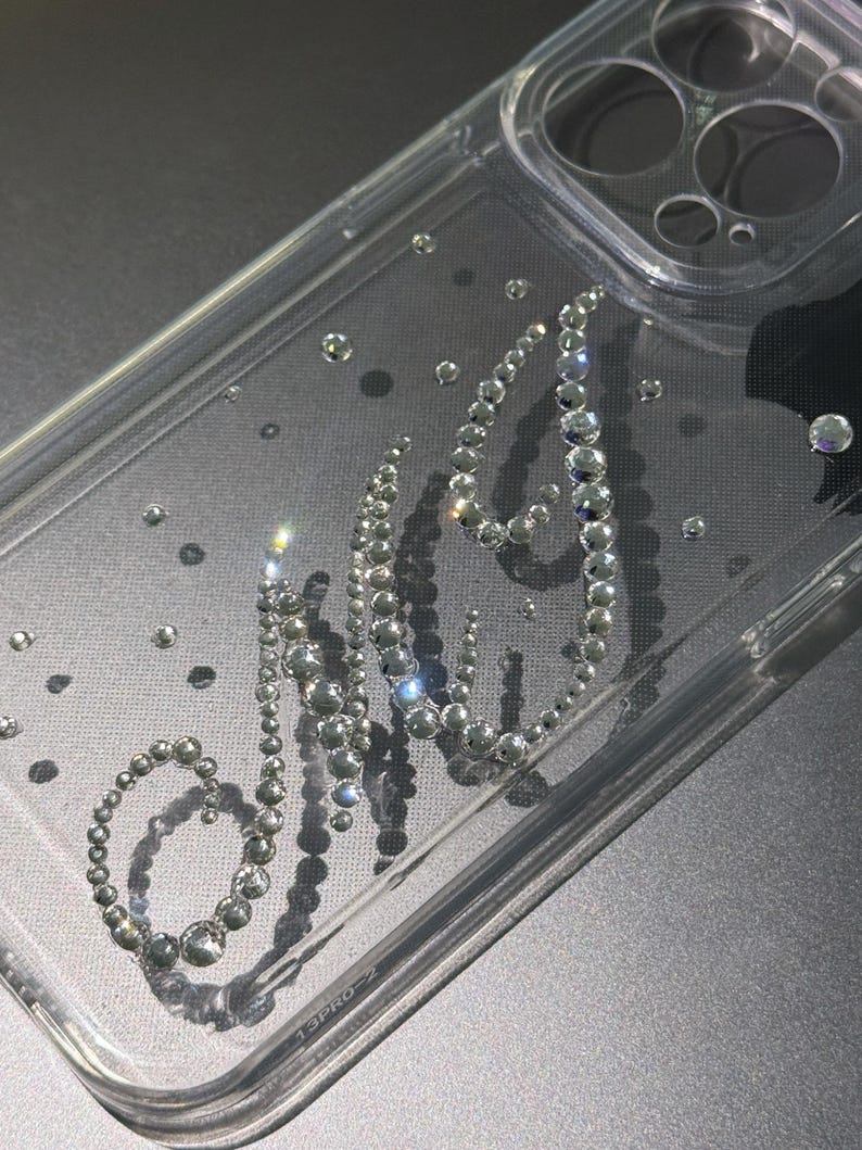 May include: Clear phone case with the word "NYC" spelled out in sparkling rhinestones. The case has a transparent design with scattered rhinestones, and is designed for a phone. The case is made of clear plastic.