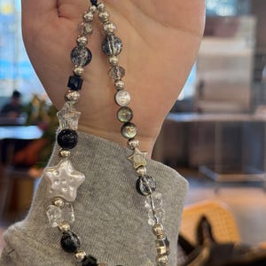 May include: A black and silver beaded phone charm with a white star charm. The charm is made with clear, black, and silver beads, and a black string.