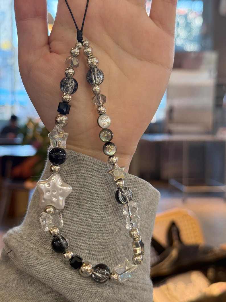 May include: A black and silver beaded phone charm with a white star charm. The charm is made with clear, black, and silver beads, and a black string.