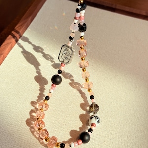 Puede incluir: Un dije de teléfono con cuentas que presenta una variedad de cuentas en rosa, negro, blanco y dorado. El dije incluye un colgante rectangular plateado con un diseño floral y una pequeña corona dorada.