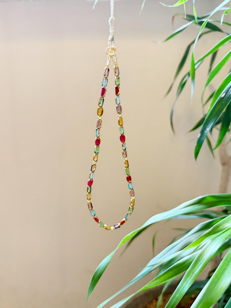 May include: A colorful beaded necklace with a gold clasp and chain. The necklace features a variety of oval-shaped beads in shades of red, blue, green, yellow, and purple. The necklace is hanging in front of a neutral background with green plants.