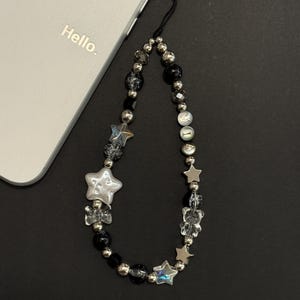 May include: A black and silver phone charm with a beaded chain and star-shaped charms. The charm is attached to a white phone case with the word "Hello" printed on it.