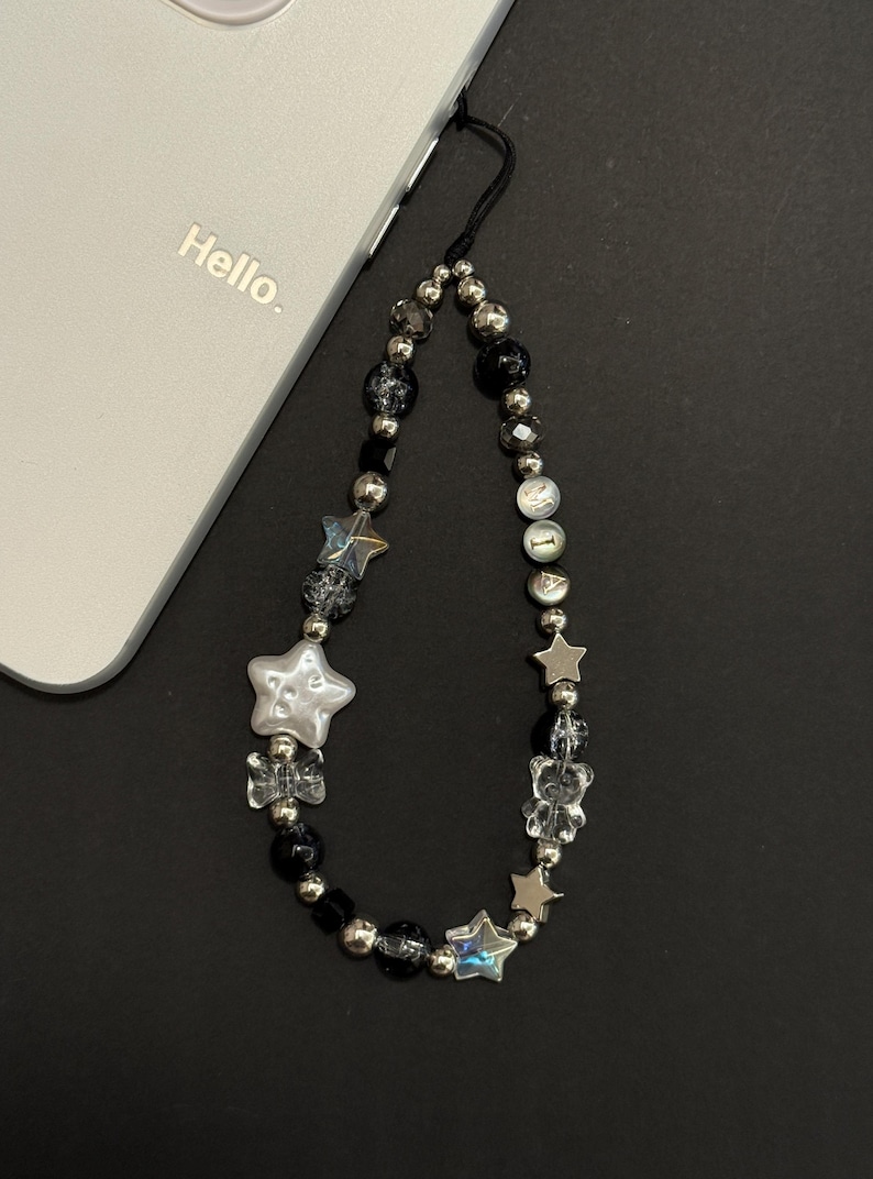 May include: A black and silver phone charm with a beaded chain and star-shaped charms. The charm is attached to a white phone case with the word "Hello" printed on it.