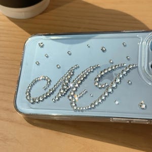 May include: Clear phone case with a light blue backing, embellished with sparkling rhinestones. The initials "MG" are spelled out in rhinestones. Additional rhinestones are scattered across the case. The case is on a wooden surface.