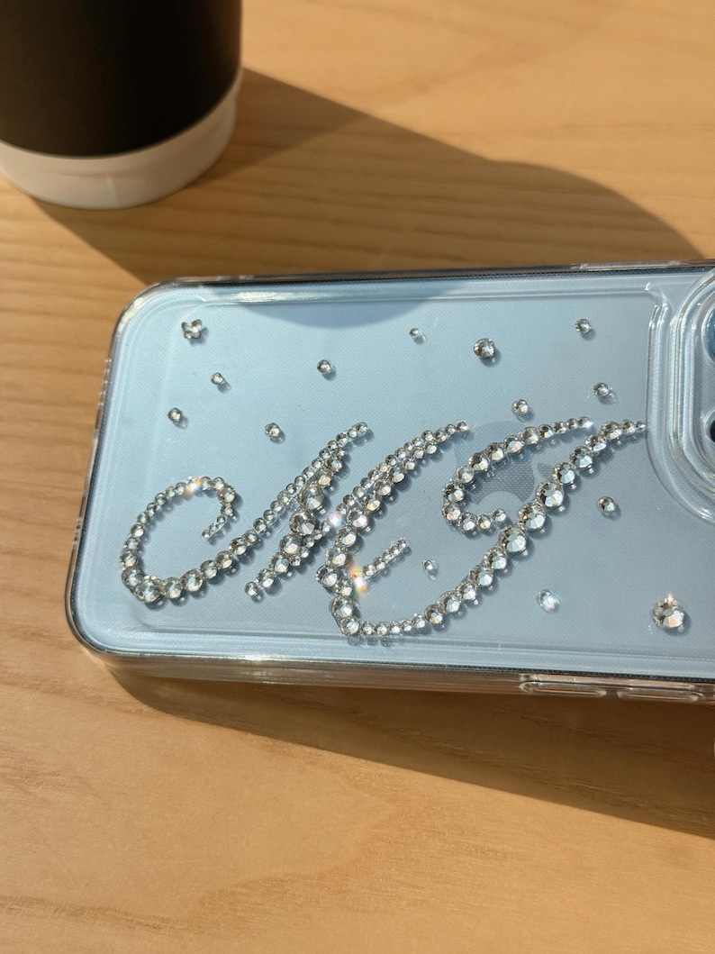 May include: Clear phone case with a light blue backing, embellished with sparkling rhinestones. The initials "MG" are spelled out in rhinestones. Additional rhinestones are scattered across the case. The case is on a wooden surface.