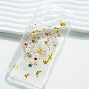 May include: Clear phone case with a space-themed design. The case is filled with gold glitter, gold stars, a planet, a rocket, and colourful gems. The case is made of clear plastic.