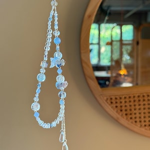 Pu&ograve; includere: Un delicato charm per telefono con perline trasparenti, azzurre e argentate. Il charm include un motivo floreale e due pendenti a forma di goccia. Lo charm &egrave; sospeso da un sottile cordino d'argento.