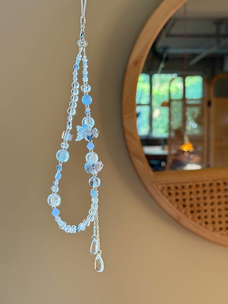 Pu&ograve; includere: Un delicato charm per telefono con perline trasparenti, azzurre e argentate. Il charm include un motivo floreale e due pendenti a forma di goccia. Lo charm &egrave; sospeso da un sottile cordino d'argento.