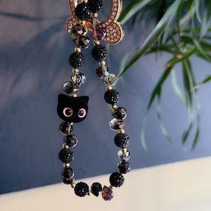May include: A black cat charm with pink eyes hangs from a gold chain with black and clear crystal beads.