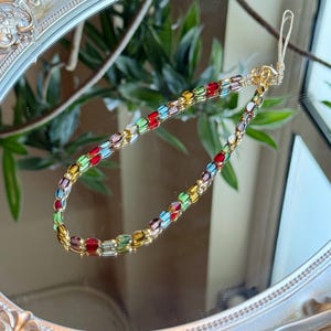 May include: A colorful beaded phone strap with gold-tone hardware. The strap features a variety of rectangular beads in red, green, yellow, and purple. The strap is reflected in a decorative, ornate mirror.