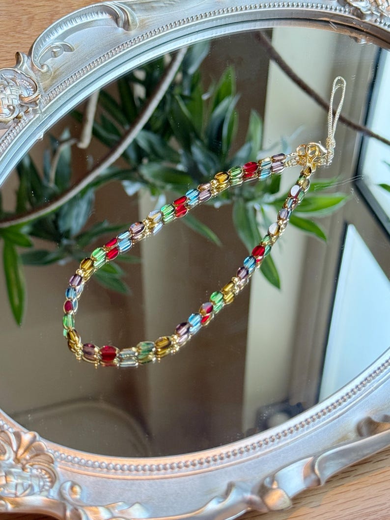 May include: A colorful beaded phone strap with gold-tone hardware. The strap features a variety of rectangular beads in red, green, yellow, and purple. The strap is reflected in a decorative, ornate mirror.