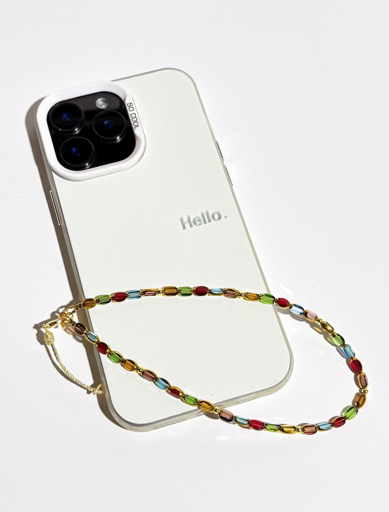 May include: A white smartphone with a protective case and the text "Hello." is shown. A colorful beaded phone strap with red, green, blue, and yellow beads is draped across the phone. The case has the text "SO COOL."