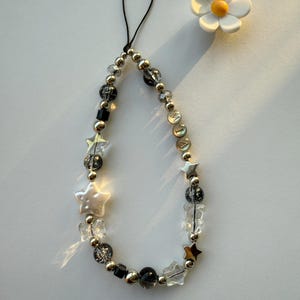 May include: A silver and black beaded phone charm with a star and crystal design. The charm has a black string loop for attaching to a phone case.