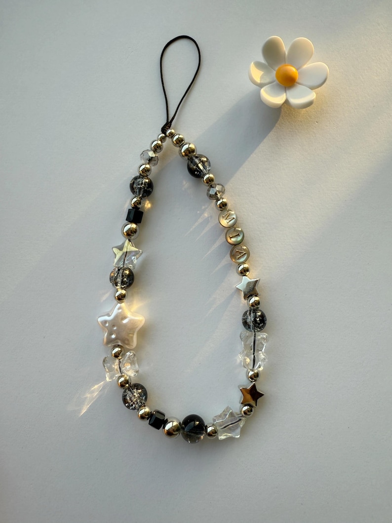 May include: A silver and black beaded phone charm with a star and crystal design. The charm has a black string loop for attaching to a phone case.