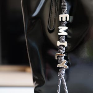 Peut inclure: Un sac fourre-tout en cuir verni noir avec un charm en corde grise et blanche épelant le nom "EMILY" en lettres argentées. Le sac a une finition brillante et une poignée argentée.