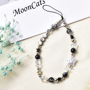 May include: A black and white beaded phone charm with a star and a white pearl charm. The charm is attached to a black string.