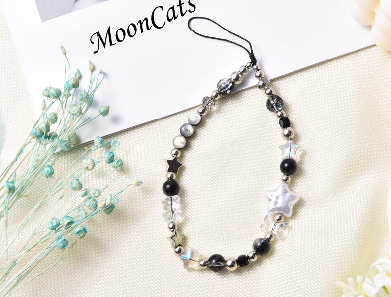 May include: A black and white beaded phone charm with a star and a white pearl charm. The charm is attached to a black string.
