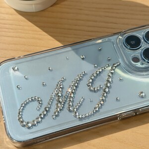 May include: Clear phone case with a light blue backing, adorned with sparkling rhinestones. The initials "MJ" are spelled out in rhinestones. Additional rhinestones are scattered across the case. The phone case is on a wooden surface.