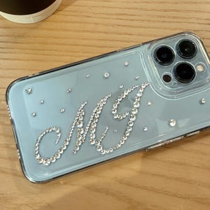 May include: A clear phone case with a light blue backing, adorned with sparkling rhinestones. The initials "MG" are spelled out in a cursive font using the same rhinestones. Additional rhinestones are scattered across the case.