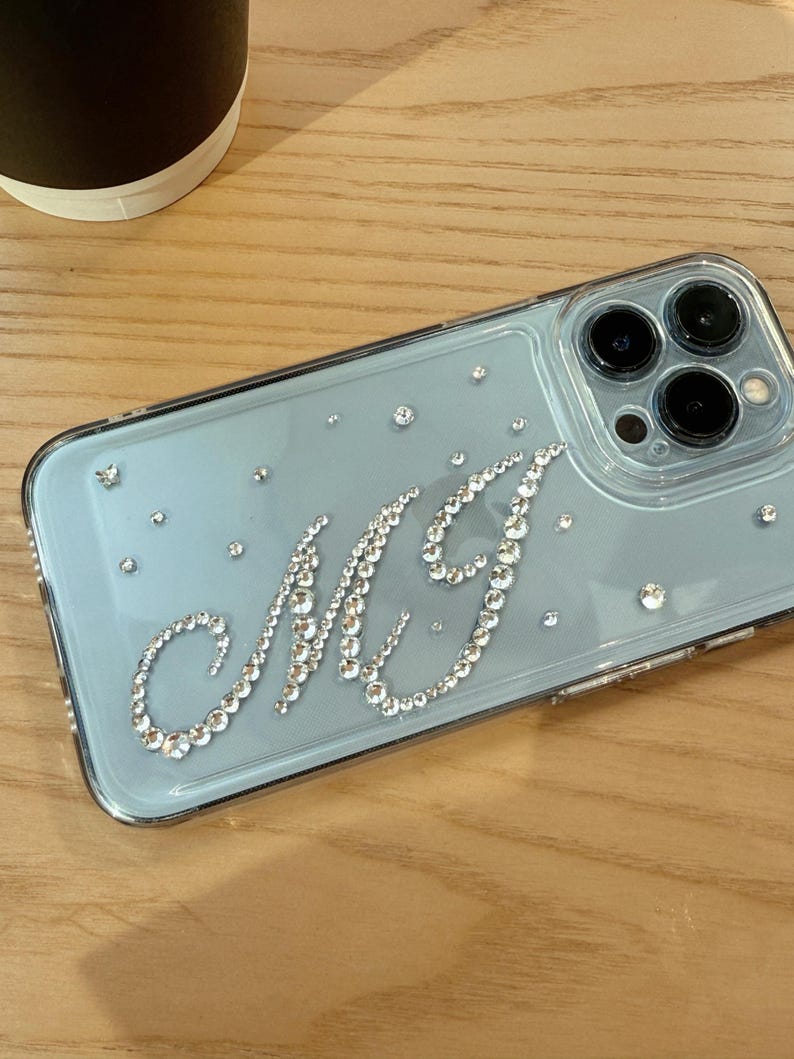 May include: A clear phone case with a light blue backing, adorned with sparkling rhinestones. The initials "MG" are spelled out in a cursive font using the same rhinestones. Additional rhinestones are scattered across the case.