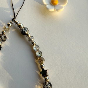 May include: A silver charm bracelet with a black string, clear and black beads, a silver star charm, and letter beads that spell "MIA". A white daisy with a yellow center is in the background.