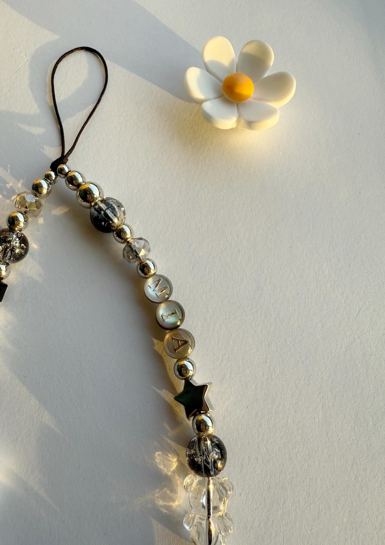 May include: A silver charm bracelet with a black string, clear and black beads, a silver star charm, and letter beads that spell "MIA". A white daisy with a yellow center is in the background.
