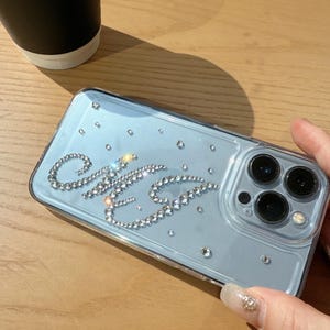 May include: Clear phone case with a light blue backing, adorned with sparkling rhinestones spelling out the initials "C&G". Additional scattered rhinestones add a touch of glamour. The case has a camera cutout and is held on a wooden surface.