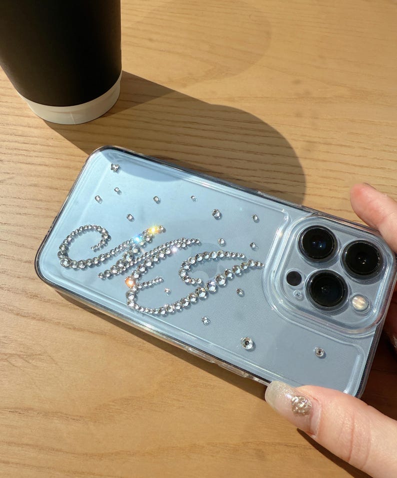May include: Clear phone case with a light blue backing, adorned with sparkling rhinestones spelling out the initials "C&G". Additional scattered rhinestones add a touch of glamour. The case has a camera cutout and is held on a wooden surface.