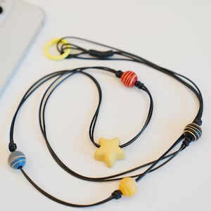 May include: A black cord phone lanyard with a yellow star charm and colorful wooden beads. The lanyard has a yellow ring and is next to a silver smartphone.