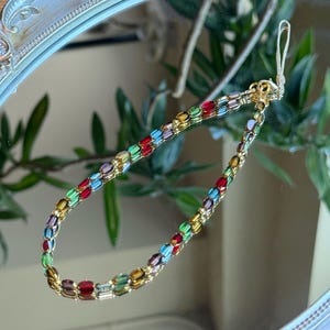 May include: A colorful beaded bracelet with gold-colored accents. The bracelet features rectangular beads in various colors, including red, green, blue, and yellow. The bracelet is displayed on a reflective surface.