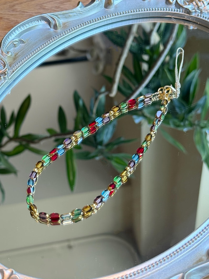 May include: A colorful beaded bracelet with gold-colored accents. The bracelet features rectangular beads in various colors, including red, green, blue, and yellow. The bracelet is displayed on a reflective surface.