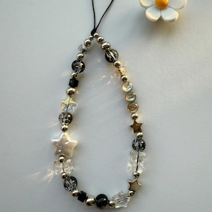 May include: A black phone charm with a silver chain and beads, including clear, black, and silver star-shaped beads. A white pearl-like star bead is in the center of the charm. A white daisy with a yellow center is in the background.