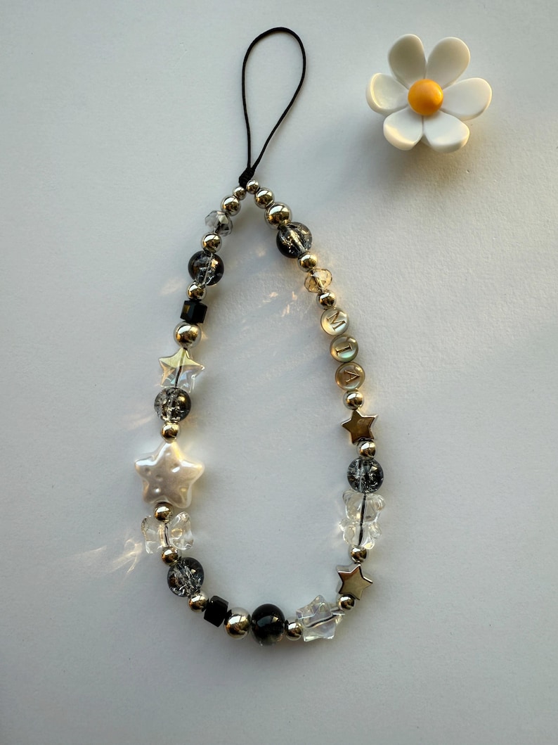 May include: A black phone charm with a silver chain and beads, including clear, black, and silver star-shaped beads. A white pearl-like star bead is in the center of the charm. A white daisy with a yellow center is in the background.