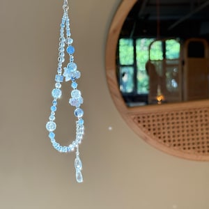 Pu&ograve; includere: Un delicato ciondolo di perline azzurre con una catena d'argento. Il ciondolo presenta varie forme di perline, tra cui disegni rotondi, sfaccettati e floreali. Il ciondolo &egrave; appeso davanti a uno specchio rotondo.