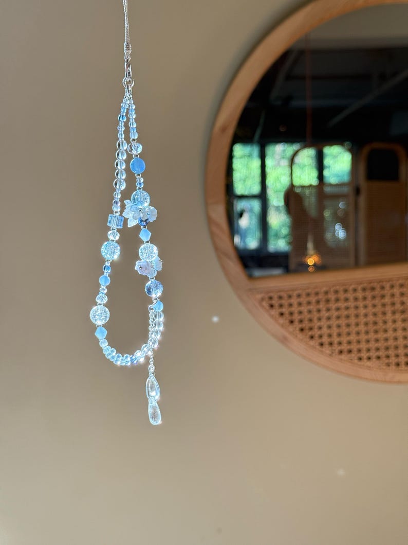 Pu&ograve; includere: Un delicato ciondolo di perline azzurre con una catena d'argento. Il ciondolo presenta varie forme di perline, tra cui disegni rotondi, sfaccettati e floreali. Il ciondolo &egrave; appeso davanti a uno specchio rotondo.