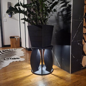 May include: A black plant stand with a round base and four legs. A black planter with a plant is on top of the stand. The stand is illuminated from below.