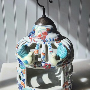 May include: A white ceramic birdcage with a dome top and a metal hanging hook. The structure features cut-out arches and is decorated with hand-painted floral patterns in orange, blue, and green, with two small sculpted birds attached.