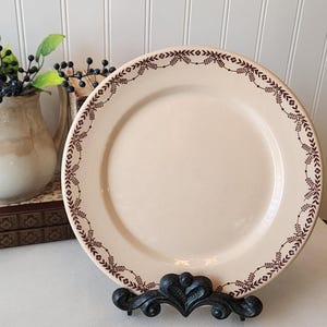 May include: A round, cream-colored ceramic plate featuring a dark brown decorative border pattern of small flowers and leafy vines. The plate is displayed upright on a black, ornate, scrolled metal stand, showcasing its detailed rim design.