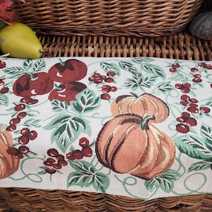 May include: A woven fabric table runner featuring a repeating pattern of orange pumpkins, deep red apples, and clusters of small red berries set against a light cream background with green leaves and thin, winding vine stems.