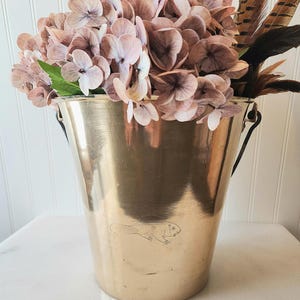 May include: A polished brass bucket featuring a small engraved beaver illustration on the front surface. The bucket is filled with a cluster of light pink hydrangea flowers and several brown and black striped feathers, with a dark metal handle attached.