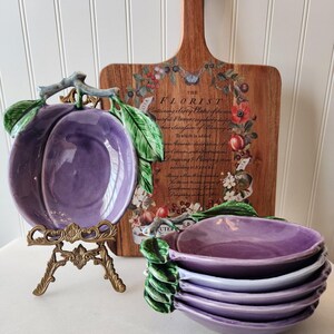 May include: A set of ceramic fruit-shaped dishes in a deep purple hue, each featuring a green leaf and stem handle. One dish is displayed upright on a gold-toned metal stand, while five others are stacked horizontally in a pile.