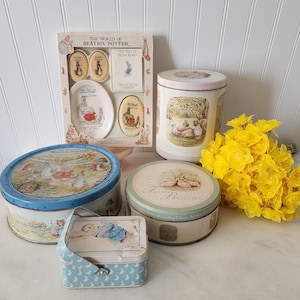 May include: A collection of Beatrix Potter themed items, including tins and a gift set. The tins are round and oval, with illustrations of Peter Rabbit and other characters. A small blue and white box with a handle is also present, along with a bouquet of yellow flowers.